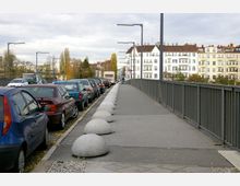 Eine städtische Brücke mit einem Fußweg rechts, gesäumt von Metallgeländern und Betonhalbkugeln als Begrenzung. Links parken Autos in einer Reihe, im Hintergrund stehen mehrstöckige Wohngebäude mit weißen und gelblichen Fassaden.