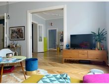 Wohnzimmer mit hellen Wänden und Parkettboden, ausgestattet mit einer Holzkommode, einem Fernseher, Pflanzen, einem kleinen Couchtisch und einem blauen Hocker. Im Hintergrund sind offene Schiebetüren erkennbar, die den Blick in einen angrenzenden Raum mit Regalen und einem gelben Möbelstück freigeben.