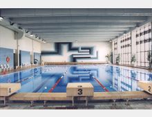 Innenansicht eines Schwimmbads mit einem rechteckigen Becken, das durch rot-weiße Leinen in Bahnen unterteilt ist. Der Raum hat helle Wände, eine blau-weiße Wandgestaltung im Hintergrund, eine Sitzbank entlang der Wand und große Fenster auf der rechten Seite, die Tageslicht hereinlassen.