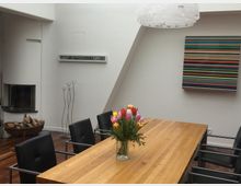 Modernes Esszimmer mit hellem Holztisch, schwarzen Lederstühlen und einer Vase mit bunten Tulpen als Tischdekoration. Im Hintergrund ein diagonales Dachfenster, ein Rundkamin mit Brennholzkorb sowie ein abstraktes Kunstwerk mit bunten Streifen an der weißen Wand.