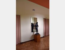 Innenraum mit einem Holzboden und einer hellen Wand, darauf eine kleine schwarze Statue auf einem Holzsockel, die vor einem rechteckigen Spiegel steht. Links und rechts befinden sich rosafarbene Vorhänge an Türöffnungen.