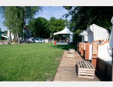Grüne Wiese mit Holzdielen am Rand, darauf Strandkörbe und Holzpaletten als Sitzgelegenheiten. Im Hintergrund stehen ein weißes Festzelt, ein Gebäude und hohe Bäume.