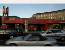 U-Bahnhof Fehrbelliner Platz in Berlin mit einem auffälligen roten Eingangsgebäude, das runde und kantige Architekturelemente kombiniert. Im Vordergrund verkehren Autos, während Passanten den Eingangsbereich betreten; im Hintergrund ist ein mehrstöckiges Gebäude mit vielen Fenstern zu sehen.