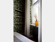 Schlafzimmer-Ecke mit floral bedruckter Tapete, einem offenen Fenster mit weißem Rahmen und halbtransparenten Vorhängen. Auf der Fensterbank steht eine gelbe Vase mit getrocknetem Gras; darunter befindet sich ein dunkler Heizkörper.