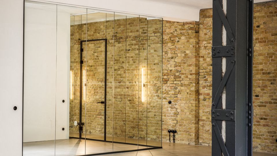 Innenraum im Loft-Stil mit freiliegender Backsteinwand, poliertem Betonboden und großer spiegelnder Schrank- bzw. Wandfläche aus Glaspaneelen. Rechts steht eine massive, genietete Stahlstütze, ergänzt durch schlichte Wandbeleuchtung und eine dunkle Tür.