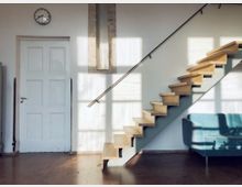 Minimalistischer Wohnraum mit einer weißen Tür, einer modernen, schwebenden Holztreppe mit Metallgeländer und einem blauen Sofa. Tageslicht fällt durch große Fenster und erzeugt Schattenmuster an der Wand.