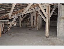 Dachboden eines alten Gebäudes mit sichtbaren Holzbalken, zum Teil stark abgenutzt, und einem offenen Dachstuhl. Der Boden ist aus Holz und teilweise beschädigt, die Wände zeigen unverputztes Mauerwerk.