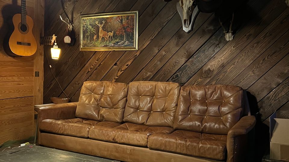 Rustikaler Aufenthaltsraum mit schräg vertäfelten Holzwänden und Holzboden, vor einer braunen Ledercouch und einem schwarzen Kistentisch. An der Wand hängen Jagdtrophäen (Geweihe und Schädel), ein Waldgemälde sowie eine Gitarre; warme Lampen beleuchten den Raum.