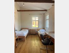 Ein schlicht eingerichtetes Zimmer mit zwei Einzelbetten aus Holz, einem kleinen Holzschrank in der Mitte und einem Schreibtisch mit Stuhl auf der rechten Seite. Die Wände sind hell, und durch ein großes Fenster fällt Tageslicht ein; sichtbare Holzbalken zieren die Decke.