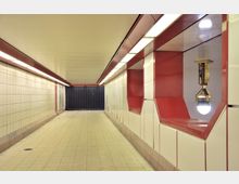 Unterirdischer Fußgängertunnel mit weißen Wandfliesen und roten Akzenten auf der rechten Seite. Die Beleuchtung erfolgt durch rechteckige Deckenleuchten, und am Ende des Tunnels befindet sich eine schwarze, geriffelte Tür oder Wand.