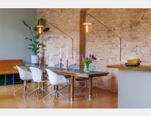 Essbereich mit Holztisch und weißen Stühlen vor einer Mauer aus Ziegelsteinen. Dekorative Details umfassen lila Tulpen in einer Vase, Kerzenhalter und moderne, goldene Wandlampen; im Hintergrund stehen eine Zimmerpflanze und eine Holzbank.