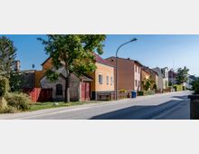 Straßenszene in einem Wohngebiet mit Einfamilienhäusern, teils aus Backstein und mit hellen Fassadenfarben. Vor den Häusern stehen Bäume, kleine Gärten und Zäune; rechts verläuft ein Gehweg entlang der asphaltierten Straße mit Straßenlaternen.