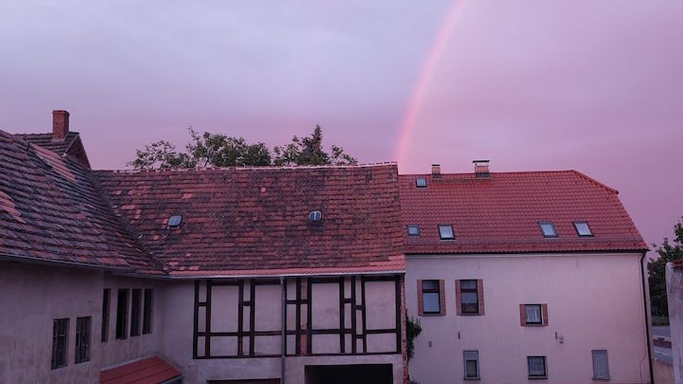 Courtyard of traditional residential buildings with red-tiled roofs, garage doors, and a central passageway, bordered by a paved area and small garden. A faint rainbow arcs across a pink evening sky above the rooftops.