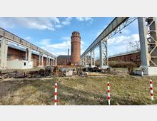 Industriegelände mit einem hohen, runden Backsteinturm im Hintergrund, flankiert von zwei langen, metallenen Tragwerkkonstruktionen. Im Vordergrund liegen verstreut alte Maschinen- und Metallteile auf einer überwachsenen Fläche.