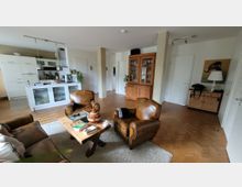 Wohnzimmer mit offener Küche und Holzboden. Im Vordergrund stehen braune Ledersessel um einen rustikalen Holztisch, im Hintergrund befinden sich eine helle Einbauküche, ein Holzvitrinenschrank und weitere Möbel.