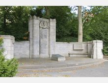 Steinerne Gedenkstätte mit Inschriftentafel, flankiert von symmetrischen Mauerelementen und Reliefverzierungen, umgeben von dichtem Grün eines Parks. Im Vordergrund ist eine geneigte Informationstafel auf einem Podest sichtbar.