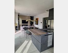 Moderne Wohnküche mit einer grauen Kücheninsel aus Steinoptik und einer Holzarbeitsplatte. Im Hintergrund ein gemütlicher Wohnbereich mit einem großen Sofa, einer wandmontierten schwarzen Fernsehanlage und dekorativen Elementen wie goldenen Statuen und einem farbigen Buddha-Bild an der Wand.