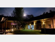 Hof im ländlichen Stil bei Nacht, umgeben von Backsteingebäuden mit großen Fenstern und Holztüren, beleuchtet durch warmes Außenlicht. Im Vordergrund stehen Tische, Stühle und ein Baum, während der Vollmond hinter Bäumen am Himmel sichtbar ist.