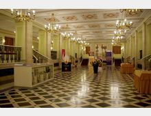 Ein elegant gestalteter Innenbereich mit Marmorwänden und hellgrünen Akzenten, verziert mit Kronleuchtern und einem schwarz-weiß karierten Boden. Im Hintergrund befinden sich Stufen, Wandgemälde mit antiken Säulenmotiven sowie Infotische und Banner.