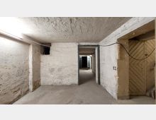 Ein leerer, heller Kellerraum mit rauen, weiß gestrichenen Wänden und einer Betonbodenfläche. Ein offener Durchgang führt in einen weiteren Bereich mit sichtbaren Holztüren, und Kabel sind entlang der Decke gespannt.