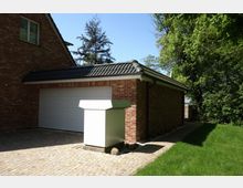 Backstein-Garage mit schwarzem Satteldach und großem weißen Garagentor, umgeben von einem gepflasterten Hof und Rasenflächen. Im Vordergrund steht ein weißer Kasten, seitlich von Bäumen und Sträuchern begrenzt.