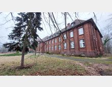 Ein großes, zweistöckiges Backsteingebäude mit weißen Fenstern und einem Satteldach, umgeben von einer Wiese und kahlen Bäumen. Im Vordergrund stehen Äste eines Nadelbaums, und ein schmaler Weg führt entlang der Gebäudeseite.