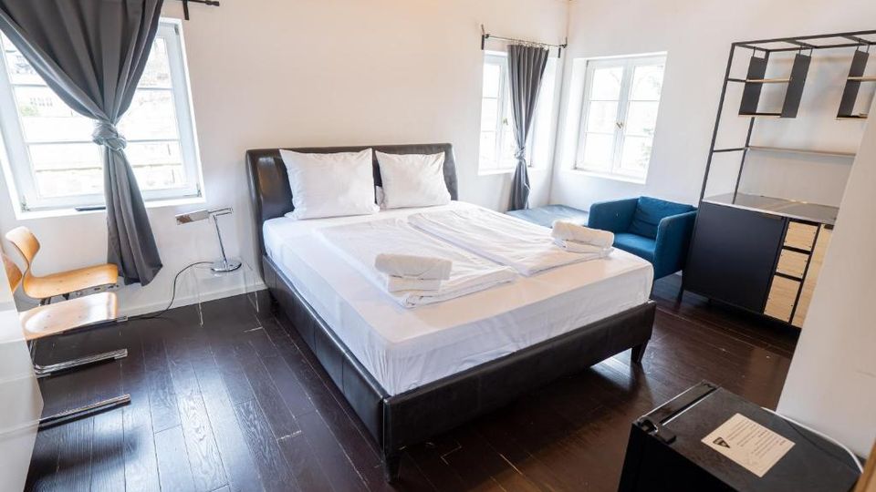 Helles Schlafzimmer mit Doppelbett, dunklem Holzfußboden und sichtbaren Deckenbalken. Drei Fenster mit grauen Vorhängen, daneben ein blaues Sofa und ein offenes Regal- und Schrankelement an der Wand.