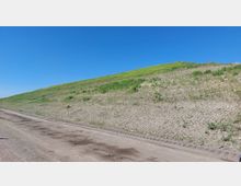 Ein unbefestigter Weg verläuft entlang einer hügeligen Fläche mit spärlichem Grasbewuchs und teils kahlem Erdreich. Der Hintergrund zeigt einen strahlend blauen Himmel.