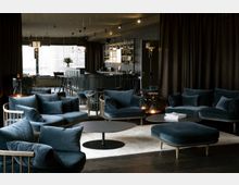 Eleganter Lounge-Bereich mit dunkelblauen Polstermöbeln, runden Beistelltischen und stilvoller Beleuchtung. Im Hintergrund befindet sich eine moderne Bar vor einer Fensterfront mit Vorhängen.