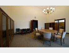 Ein Besprechungsraum mit einem runden Holztisch und sechs gepolsterten Stühlen. An den Wänden stehen hohe Schränke mit Glastüren und offene Regale; eine Deckenleuchte mit mehreren Lampen hängt in der Mitte.