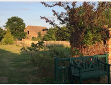 Grüne Wiese mit einem alten Backsteinzaun und einer grünen Holzbank im Vordergrund, umgeben von Bäumen und Sträuchern; im Hintergrund ein rustikales Gebäude mit Ziegeldach und ein Holzzaun.