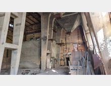 Innenansicht eines verlassenen Industriebauwerks mit hohen Betonstützen, rohen Backsteinwänden und sichtbaren Rohren sowie Kabeln an der Decke. Der Raum enthält ungenutzte Holzelemente und diverse Metallstrukturen, und natürliches Licht fällt durch Fenster und offene Bereiche.