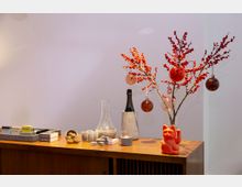 Holzkommode mit dekorativen Gegenständen, darunter ein roter Glückskatze-Figur, ein gläserner Krug, eine Flasche in einem Steinhalter, Nüsse in einer kleinen Schale und Zweige mit roten Beeren und Glaskugeln. Der Hintergrund ist eine weiße Wand.