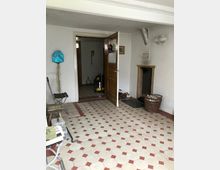 Ein einfacher Innenraum mit rot-weiß gemusterten Fliesenboden, einer geöffneten Holztür, die in einen weiteren Raum führt, einer kleinen Kamintür an der Wand und einem geflochtenen Korb daneben. Einige Werkzeuge und Utensilien stehen verstreut auf dem Boden und einer Sitzgelegenheit.