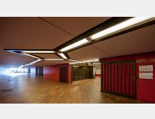 Unterirdischer U-Bahn-Bereich mit gelb-braunen Fliesen, rot verkleideten Wänden und modernem Deckenlampen-Design. Im Bild sind ein Treppenabgang, Hinweisschilder zu den Linien U5 und U2 sowie Aushänge mit Fahrplänen zu sehen.