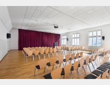 Ein moderner Veranstaltungsraum mit Holzfußboden, einer Bühne mit rotem Vorhang und mehreren Reihen von Holzstühlen mit schwarzer Polsterung. Große Fenster lassen Tageslicht herein, und an den Wänden befinden sich Lautsprecher.