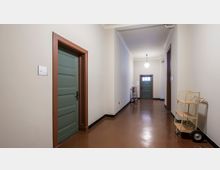 Flur mit hellen Wänden und braunem Boden; an den Seiten befinden sich grüne Türen mit braunen Türrahmen. Im Hintergrund sind ein Rollwagen und ein Regal mit Rollen zu sehen, die auf dem glänzenden Boden stehen, und eine hängende Deckenlampe leuchtet den Bereich aus.