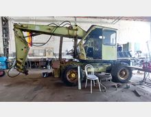 Ein alter, gelb-grüner Radbagger in einer Werkstatthalle mit weiß gemauerten Wänden. Im Hintergrund sind Werkbänke, Werkzeuge und eine Deutschlandfahne sichtbar.