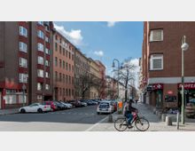Straßenszene in einer Stadt mit mehrstöckigen Wohn- und Geschäftshäusern aus Backstein und verputztem Mauerwerk, teils im Stil der Gründerzeit. Geparkte Autos stehen entlang der Straße, ein Radfahrer fährt auf dem Gehweg, und im Vordergrund sind Laternen und ein Briefkasten zu sehen.