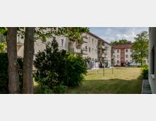 Mehrfamilienhäuser mit drei Stockwerken stehen an einer schmalen Grünfläche, die von Wäscheleinen und vereinzelten Büschen sowie Bäumen gesäumt ist. Im Hintergrund sind weitere Gebäude und geparkte Autos zu sehen.