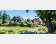 Ruhige Wohnstraße mit Einfamilienhäusern und Vorgärten, umgeben von Bäumen und Hecken. Eine asphaltierte Geh- und Fahrbahn führt durch die grüne Umgebung, rechts steht ein großer Baum im Vordergrund.