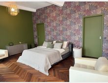 Schlafzimmer mit modernem Design: Ein großes Bett mit grauer Bettdecke und mehreren Kissen steht vor einer Wand mit floraler Tapete in Lila- und Beigetönen. Links daneben befindet sich eine grüne Wand mit einer Kommode, rechts zwei grüne Türen, ein weißer Sessel und ein Holzboden im Fischgrätmuster bestimmen den Raum.