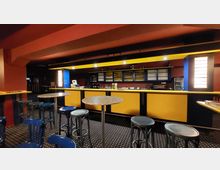 Eine Bar mit einer gelb-schwarzen Theke, darüber ein Regal mit Gläsern und Flaschen. Vor der Theke stehen runde Tische und blaue Hocker auf einem Teppichboden mit Punktmuster, umgeben von rot gestrichenen Wänden.