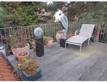 Terrasse mit Steinboden, umgeben von einem Metallgeländer, dekoriert mit Pflanzentöpfen und zwei kleinen Skulpturen. Im Hintergrund sind hohe Bäume zu sehen, und eine Holzliege steht auf der rechten Seite.