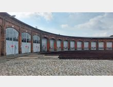 Halbrundes historisches Bahnbetriebswerk mit roten Backsteinwänden, großen Toren und vereinzelten Fenstern. Im Vordergrund verlaufen Schienen und der gepflasterte Hofbereich.