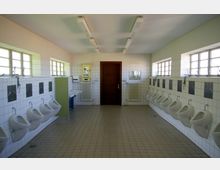 Öffentliche Toilette mit gefliesten Wänden und Boden, mehreren Urinalen an den Seitenwänden sowie einer Holztür in der Mitte der Rückwand. An der Wand befinden sich Fenster mit Glasbausteinen, Spiegel, Waschbecken und Heizkörper.