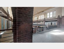 Innenraum eines historischen Gebäudes mit roten Backsteinwänden, hohen Fenstern mit Sprossen und metallenen Geländern. Auf beiden Seiten befinden sich Reihen von Türen, die zu kleinen Räumen oder Zellen führen, und die Etage wird durch eine Galerie verbunden.