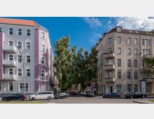 Straßenecke in einer Wohngegend mit zwei mehrstöckigen Häusern, eines in rosa und das andere in beige, beide mit Balkonen; dazwischen stehen Bäume, und am Straßenrand parken mehrere Autos.