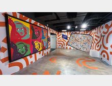 Innenraum eines modernen Museums mit bunten, abstrakten Kunstwerken. An den Wänden hängen ein großer Pop-Art-Druck mit wiederholenden Porträts sowie ein schräg montiertes Gemälde mit chaotischen Farbspritzern, umgeben von orange-weißen Wandmustern.