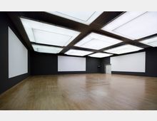 Großer, leerer Raum mit schwarzwandigen Wänden und hellem Holzfußboden; an der Decke befinden sich rechteckige, leuchtende Elemente in einem Gitterraster. Weiße Leinwände bedecken Abschnitte der Wände und eine geschlossene Tür ist an der rechten Seite zu sehen.